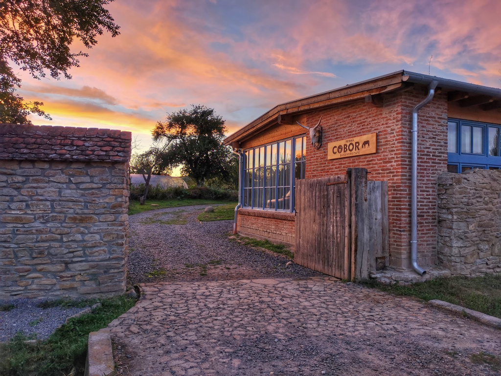 image of Cobor biodiversity farm showing the seminar building, the farm logo and a stone wall plus a wooden gate during sunset
